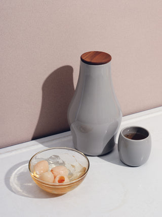 A grey carafe, small ceramic cup, and glass bowl with small pale pink fruits on a white table.