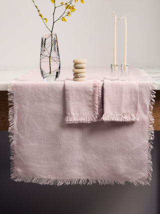 A table with a blush linen runner and 2 blush linen napkins folded on top. A glass vase with yellow flowers, a pile of white stones and 2 candle sticks sits on top of the linens.