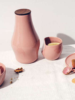 A rose carafe with a wood lid next to a curved ceramic cup with a lemon on a white table cloth.