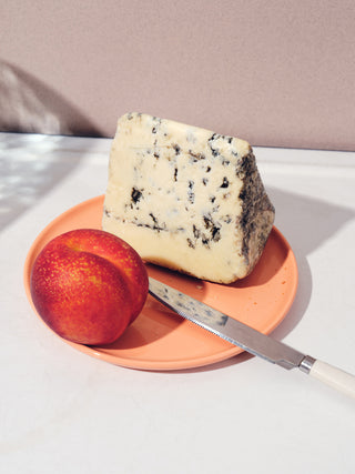 A peach, wedge of blue cheese, and butter knife on a flat coral plate on a white table with a mauve background
