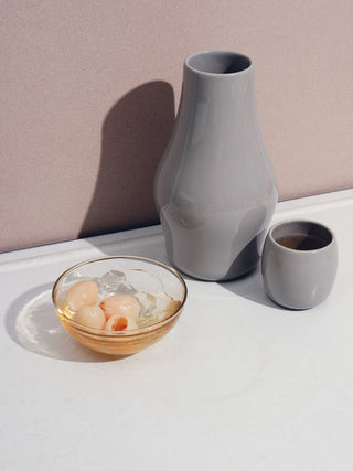 A grey carafe, small ceramic cup, and glass bowl with small pale pink fruits on a white table.