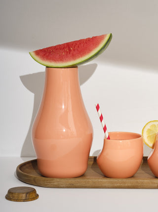 One coral carafe with a watermelon slice balanced on top next to one coral medium cup with a straw on a medium tray