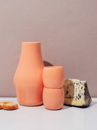 Two small coral ceramic cups stacked next to a coral carafe and wedge of blue cheese on a white table and mauve background.