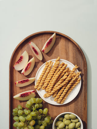 Oval wooden tray filled with peach slices, bread twists, olives, and grapes
