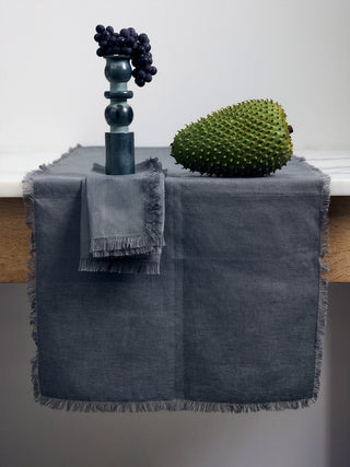 A table with a slate linen runner and a slate linen napkin. A sculpture balancing grapes sits on top while a durian fruit sits to the side.