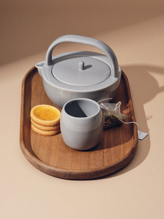 Small grey teapot with a small grey cup next to biscuits and a teabag on top of a medium tray