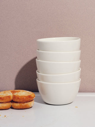 A stack of five small white bowls next to two stacked cookies on a white table with a mauve background