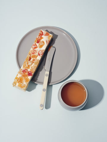 Grey ceramic plate with a slice of fruit bread, knife, and small matching cup beside