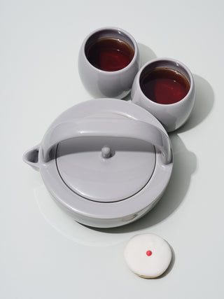 Grey ceramic teapot next to two short grey cups and a cupcake